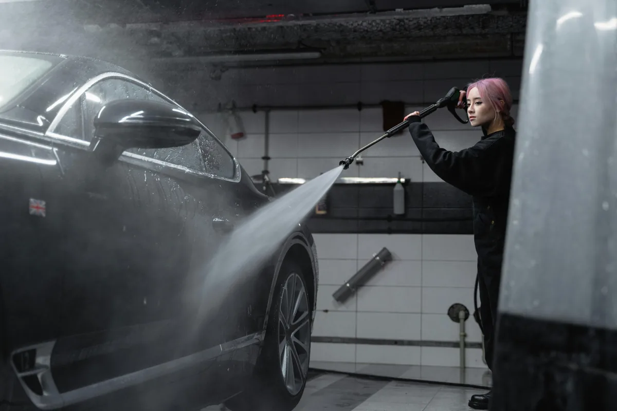 Person pressure washing dark vehicle in indoor garage cleaning facility