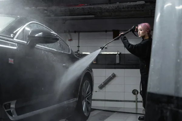 Person pressure washing dark vehicle in indoor garage cleaning facility