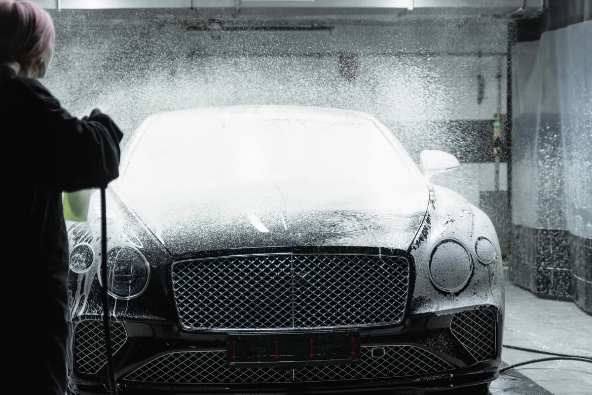 Person spraying high-pressure water to clean luxury car exterior at wash station