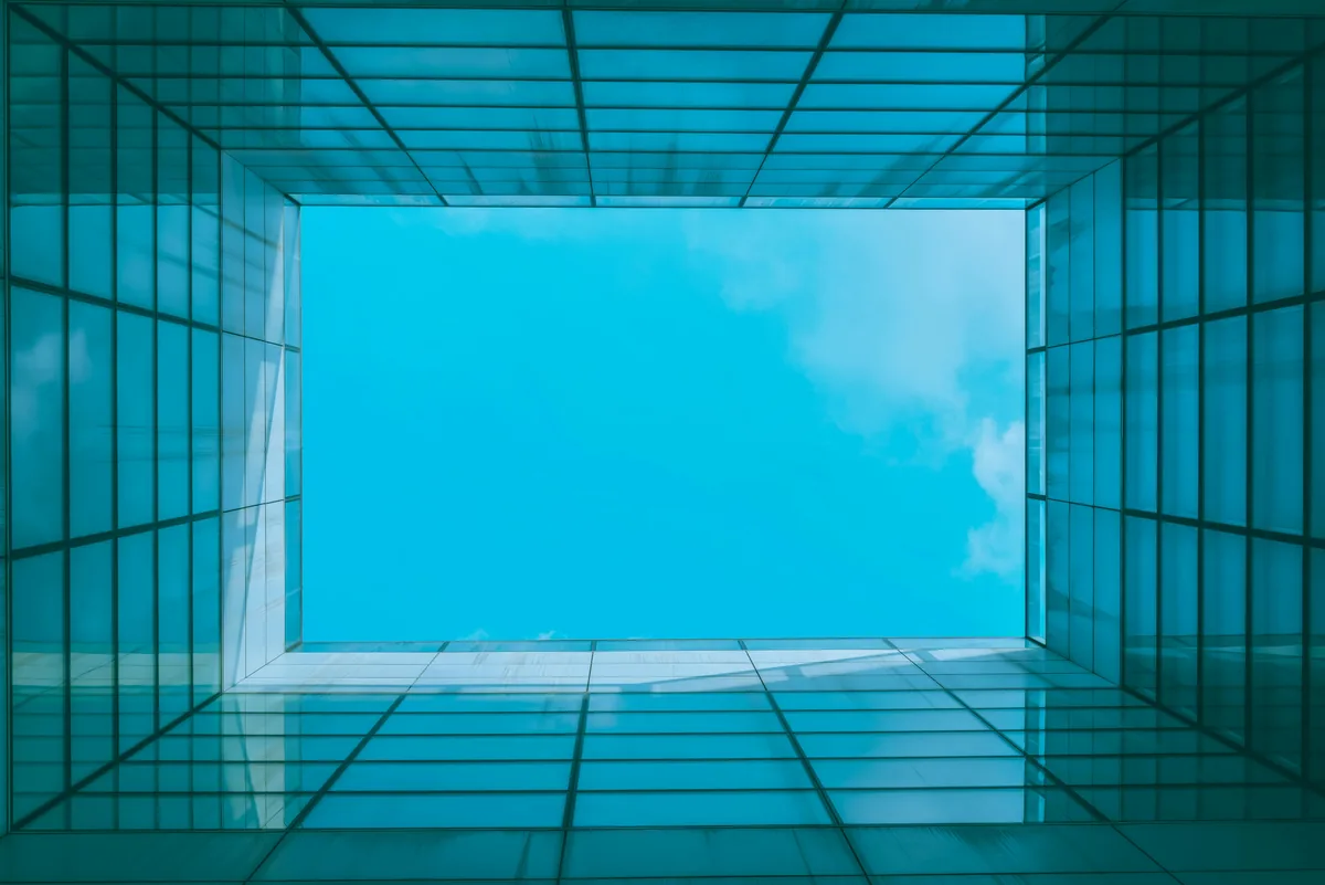 Upward view through glass building facade toward blue sky and clouds