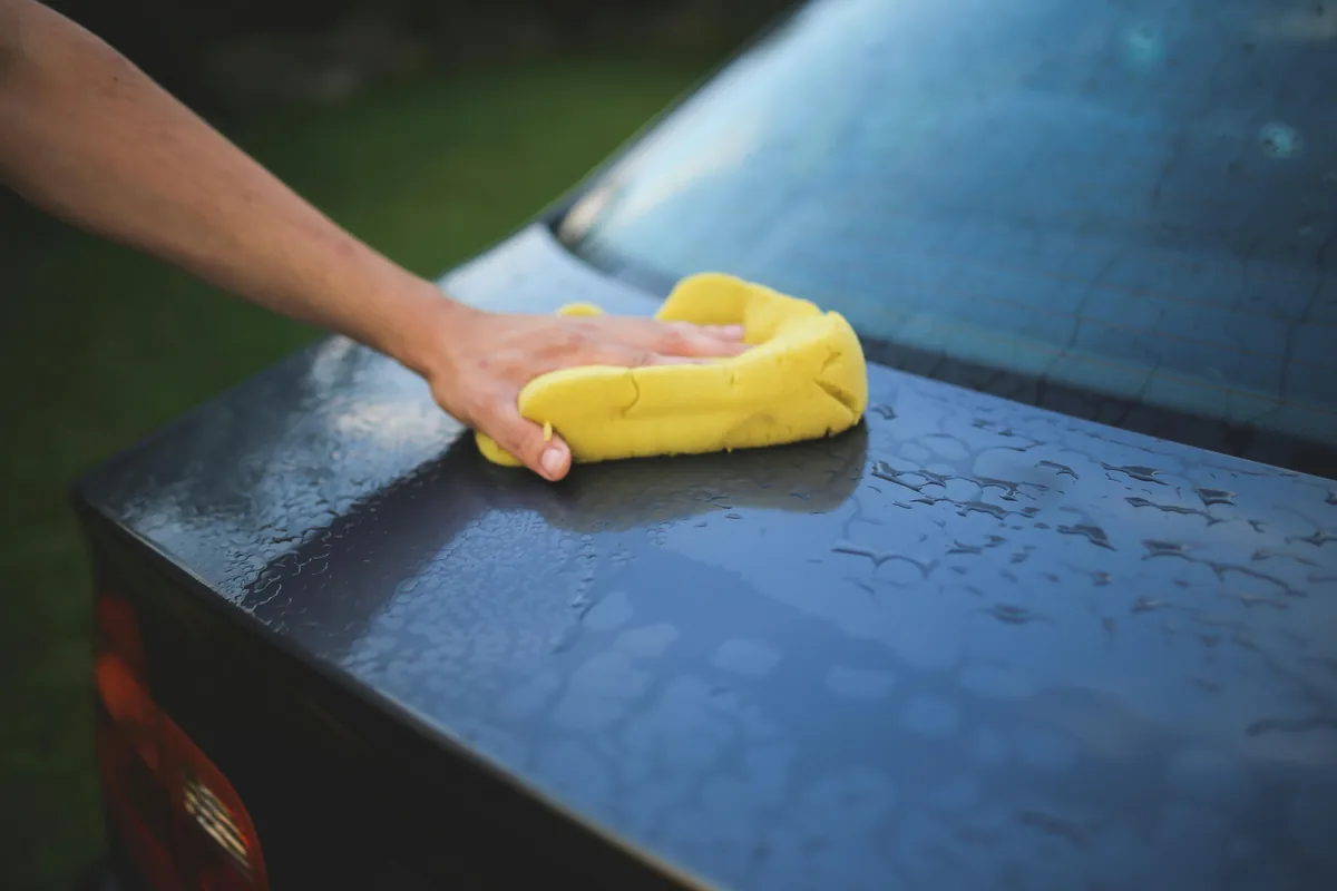 Person wiping wet blue car hood with yellow microfiber cloth outdoors