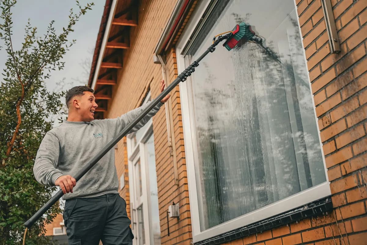 Worker using long-reach cleaning tool to wash large residential window on brick home